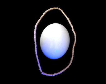 May include: A white egg sits inside a circle of light brown beads against a black background.