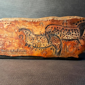Peut inclure: Plaque rectangulaire en céramique représentant des peintures rupestres de chevaux et d'empreintes de mains en marron et noir. Le texte "Grotte du Pech-Merle la frise des chevaux Ponctués" est visible. L'œuvre rappelle l'art préhistorique.
