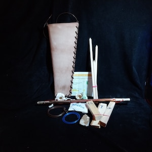 May include: A leather quiver with a brown strap, three wooden arrows, a small bag of fire starter, a piece of flint, a small piece of wood, and a piece of paper with text.