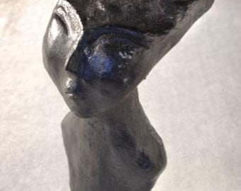 May include: A dark gray sculpture of a stylized human head and upper torso. The sculpture has a smooth, reflective surface and features a prominent brow, a defined nose, and a small mouth. The figure's head is tilted slightly upward.