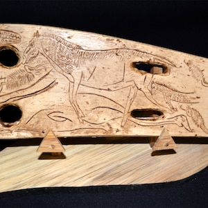 Pode incluir: Uma placa de madeira esculpida que representa uma cena de animais, possivelmente cavalos ou veados, com detalhes intrincados. A placa é de cor castanho claro e é exibida num suporte de madeira.