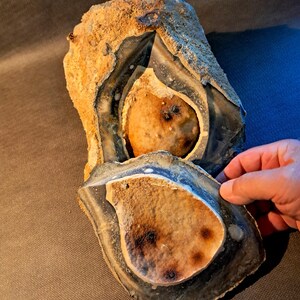 Könnte beinhalten: Ein großer, verwitterter Feuerstein mit geschichtetem Aussehen. Der Stein hat eine raue, strukturierte Oberfläche mit Schattierungen von Braun, Beige und Grau. Das Innere zeigt einen helleren, abgerundeten Kern. Eine Hand hält ein Stück des Steins.