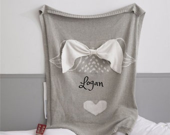 May include: A gray knitted baby blanket with a white bow and heart design. The blanket is personalized with the name "Logan".