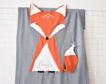 May include: A gray knitted baby blanket with a large orange fox applique. The fox has a white belly and a white patch on its head. The blanket is personalized with the name "Logan".