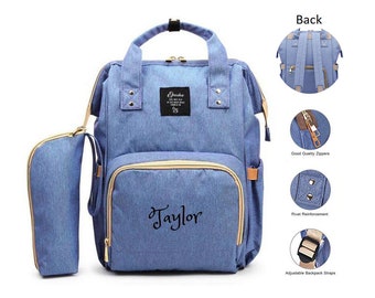 May include: A blue denim diaper bag with a personalized name "Taylor" embroidered on the front. The bag has a zippered pocket on the front and two side pockets. It also comes with a matching blue denim pencil case.
