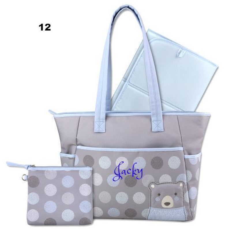 May include: A gray and blue diaper bag with a polka dot pattern and a bear applique. The bag has a large main compartment and a smaller zippered pocket. The bag also includes a matching changing pad. The bag is personalized with the name "Jacky".