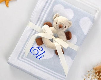 May include: A light blue baby blanket with white hearts and a white border. The blanket is tied with a white ribbon and has a brown and white teddy bear rattle attached. The blanket is personalized with the name "Eli" in blue.