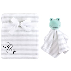 May include: A white and gray striped baby blanket with a white satin ribbon tied around it. The blanket is personalized with the name "Alex" in black cursive lettering. A plush green frog toy is attached to a smaller white and gray striped blanket.