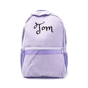 Pode incluir: Uma mochila de seersucker lavanda com o nome "Tom" em escrita preta. A mochila tem um compartimento principal com zíper, um bolso frontal e bolsos laterais de malha. O design apresenta listras verticais.