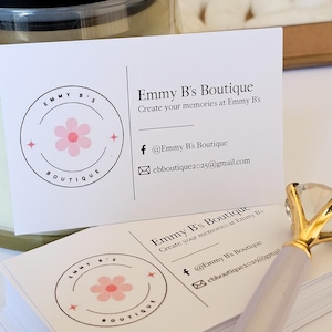 K&ouml;nnte beinhalten: Ein Stapel wei&szlig;er rechteckiger Visitenkarten f&uuml;r Emmy B's Boutique mit einem rosa Blumenlogo, schwarzem Text und Kontaktdaten. Ein Stift mit einem gro&szlig;en, klaren, diamantf&ouml;rmigen Aufsatz und einem goldfarbenen Metallsockel liegt auf den Karten.