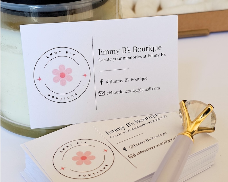 K&ouml;nnte beinhalten: Ein Stapel wei&szlig;er rechteckiger Visitenkarten f&uuml;r Emmy B's Boutique mit einem rosa Blumenlogo, schwarzem Text und Kontaktdaten. Ein Stift mit einem gro&szlig;en, klaren, diamantf&ouml;rmigen Aufsatz und einem goldfarbenen Metallsockel liegt auf den Karten.