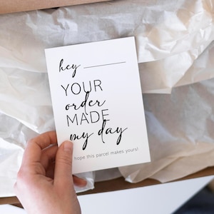 May include: A white card with black text that reads "hey ____ YOUR order MADE my day" is held in a person's hand. The card is inside a cardboard box with white tissue paper. The text "hope this parcel makes yours!" is at the bottom.