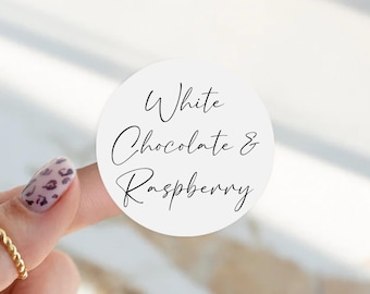 Könnte beinhalten: Ein runder weißer Aufkleber mit dem schwarzen Text White Chocolate & Raspberry in einer kursiven Schriftart. Der Aufkleber wird von einem Finger gehalten und zeigt den aufgedruckten Geschmacksnamen deutlich auf dem weißen Hintergrund.