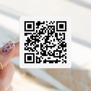 May include: A square white sticker with a black QR code. The code is composed of black squares and rectangles arranged in a complex pattern. The sticker is held by a hand with a gold ring and a patterned nail.