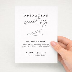 Könnte beinhalten: Eine weiße rechteckige Karte mit schwarzem Text, der OPERATION secret peg lautet, gefolgt von einer Strichzeichnung einer Wäscheklammer mit kleinen Sternen. Darunter steht YOUR SECRET MISSION: Put a peg on as many guests as possible without getting caught! OLIVIA and JAMES 9TH SEPTEMBER 2029.