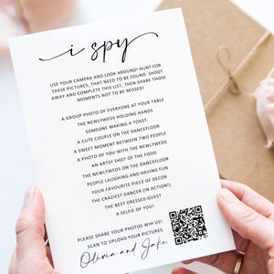 May include: A white rectangular wedding I Spy game card with black text and a QR code. The card lists photo prompts for guests, including a group photo, the newlyweds, and dancing. The bottom features the names Olivia and Jake in script font.