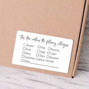 May include: A rectangular white sticker with rounded corners featuring black text that reads, This item contains the following allergens: followed by a checklist of options including Gluten, Milk, Peanuts, Nuts, Eggs, Lupin, Dairy, Soya, Sesame, Sulphites/ Sulphur Dioxide, and Other.