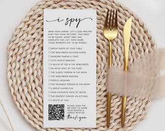 May include: A white rectangular wedding I Spy game card with black text and a QR code, featuring a list of photo prompts. The card is positioned next to a gold fork and a gold knife, which are tied together with a sheer light beige ribbon.