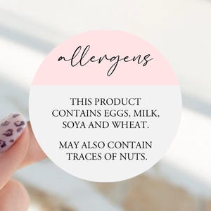 May include: A round sticker with a light pink top section featuring the word allergens in black script. The white bottom section contains the black text: THIS PRODUCT CONTAINS EGGS, MILK, SOYA AND WHEAT. MAY ALSO CONTAIN TRACES OF NUTS.