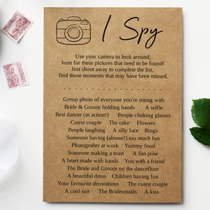 May include: A rectangular brown card featuring a black line drawing of a camera and the text I Spy. Below, instructions and a list of wedding-themed photo prompts are printed in black ink, including items like group photo, bride and groom, and cake.