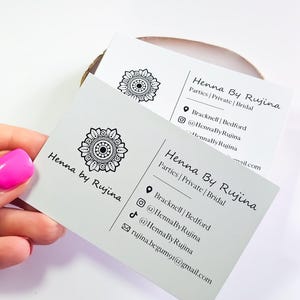 K&ouml;nnte beinhalten: Zwei wei&szlig;e rechteckige Visitenkarten mit einem schwarzen Mandala-Logo und dem Text Henna By Rujina. Die Karten f&uuml;hren die Dienstleistungen Parties, Private und Bridal auf sowie Standortdetails f&uuml;r Bracknell und Bedford, Social-Media-Profile und eine E-Mail-Adresse.