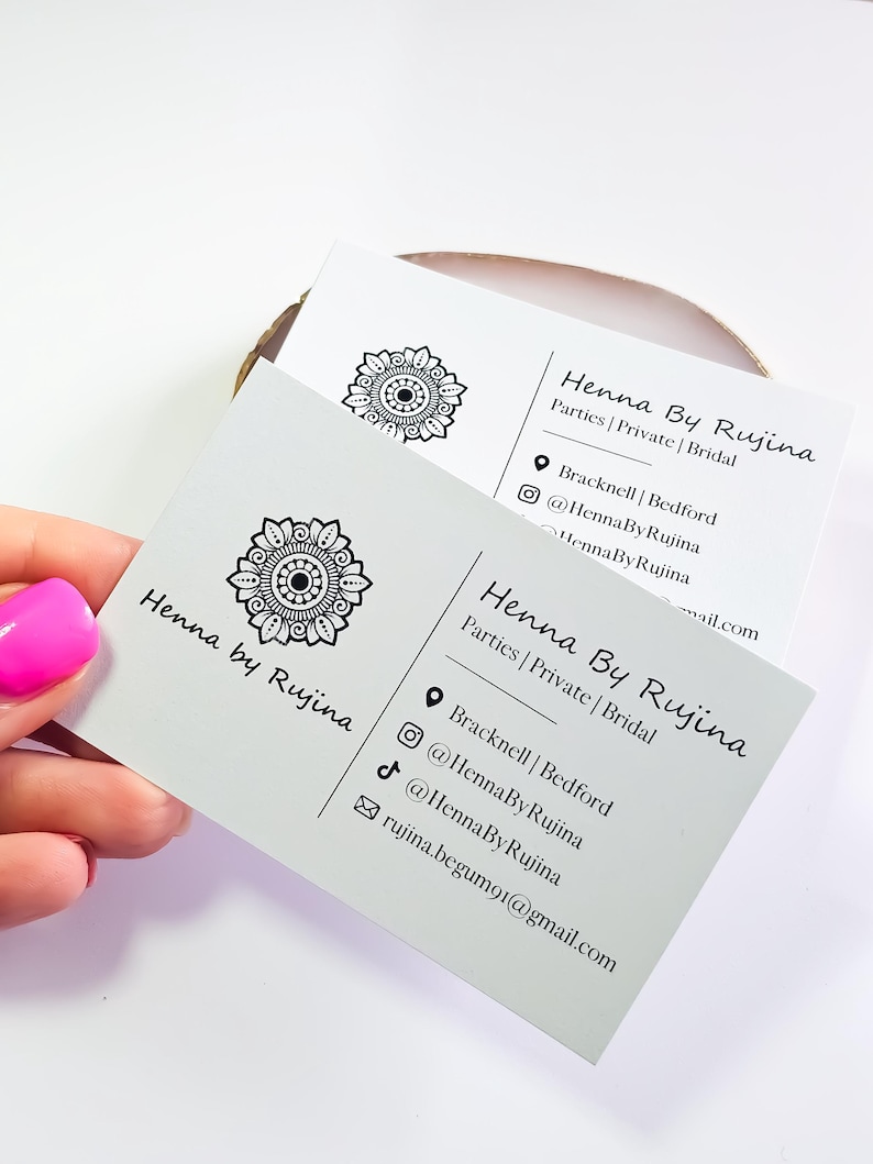 K&ouml;nnte beinhalten: Zwei wei&szlig;e rechteckige Visitenkarten mit einem schwarzen Mandala-Logo und dem Text Henna By Rujina. Die Karten f&uuml;hren die Dienstleistungen Parties, Private und Bridal auf sowie Standortdetails f&uuml;r Bracknell und Bedford, Social-Media-Profile und eine E-Mail-Adresse.
