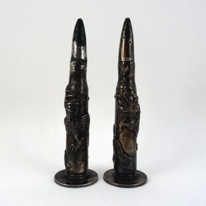 May include: Two silver-colored salt and pepper shakers. One is a solid bullet shape, the other has holes on top. Both are decorated with a dragon design. The shakers have a round base and are set against a white background.