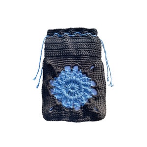 Può includere: Una borsa a coulisse all'uncinetto grigio scuro con un motivo floreale blu. La borsa presenta un fiore blu al centro e cordini blu. La borsa è realizzata con un filato spesso e strutturato.