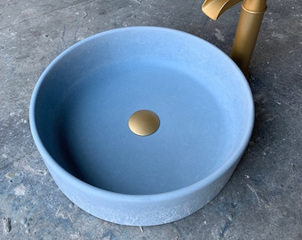 May include: A light blue round ceramic sink with a gold drain stopper. The sink is on a gray concrete surface. A gold faucet is visible in the background.