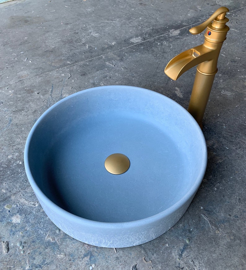 May include: A light blue round ceramic sink with a gold drain stopper. The sink is on a gray concrete surface. A gold faucet is visible in the background.