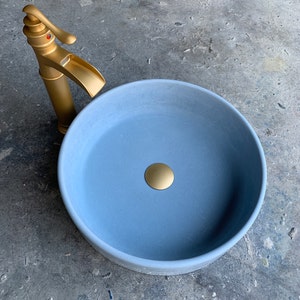 May include: A light blue ceramic bathroom sink with a gold drain stopper. The sink is round and has a smooth, matte finish. A gold faucet is visible in the background.