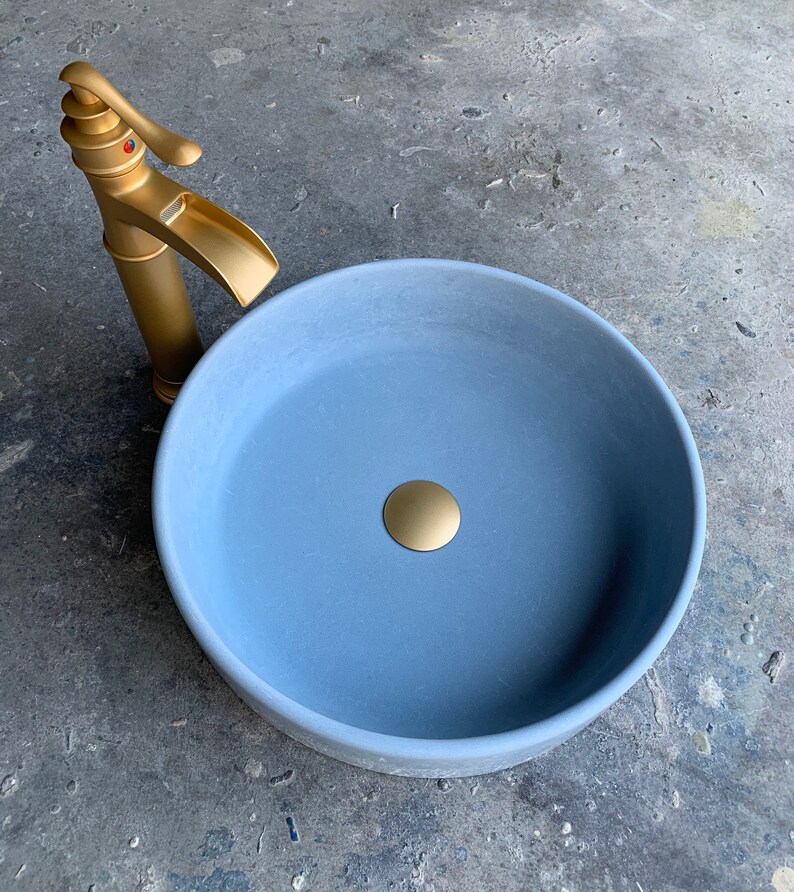May include: A light blue ceramic bathroom sink with a gold drain stopper. The sink is round and has a smooth, matte finish. A gold faucet is visible in the background.