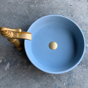 May include: A light blue ceramic bathroom sink with a gold faucet and drain stopper.