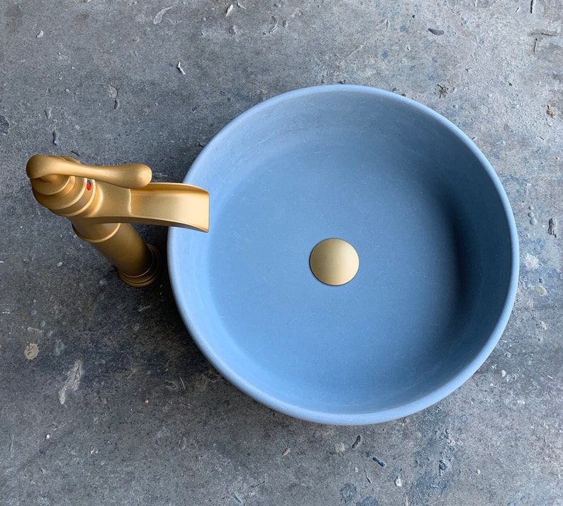 May include: A light blue ceramic bathroom sink with a gold faucet and drain stopper.