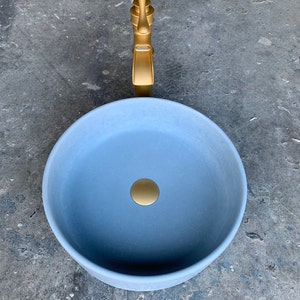 May include: A round, light blue ceramic sink with a gold drain stopper and a gold faucet. The sink is on a gray concrete surface.