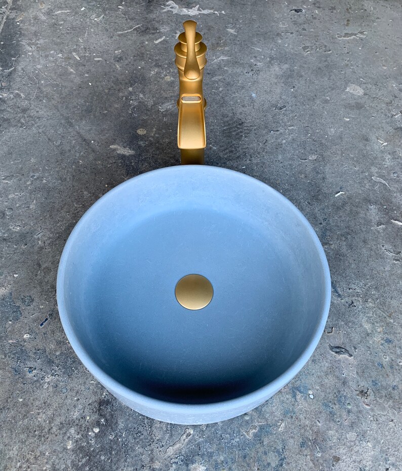 May include: A round, light blue ceramic sink with a gold drain stopper and a gold faucet. The sink is on a gray concrete surface.
