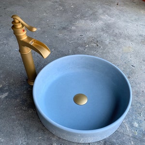 May include: A light blue ceramic sink with a gold faucet and drain stopper. The sink is round and has a smooth, matte finish.