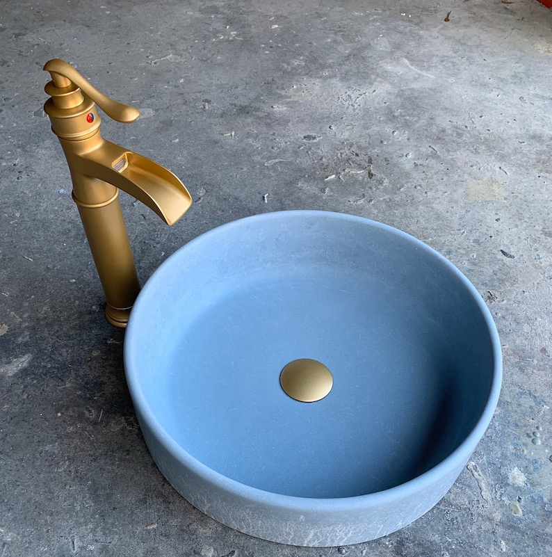 May include: A light blue ceramic sink with a gold faucet and drain stopper. The sink is round and has a smooth, matte finish.