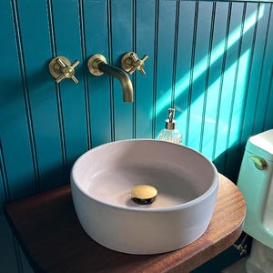 May include: A white ceramic vessel sink with a gold drain stopper sits on a dark wood countertop. The sink is mounted on a teal blue wall with gold hardware.