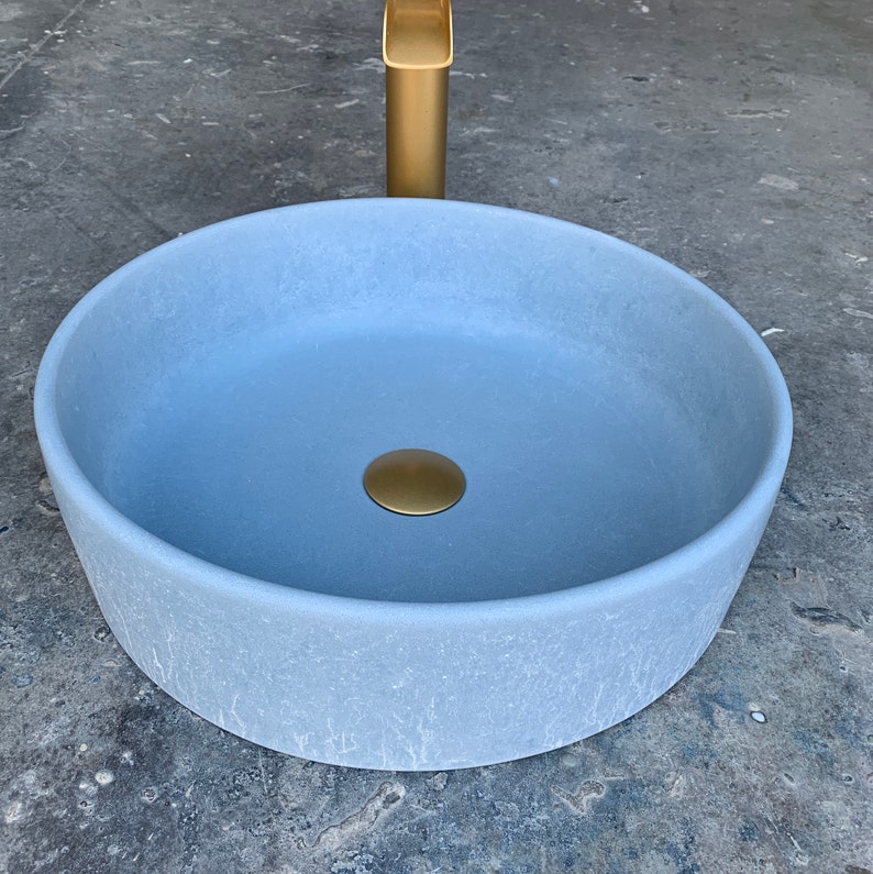 May include: A round, light blue ceramic bathroom sink with a gold drain stopper. The sink is on a gray concrete surface.