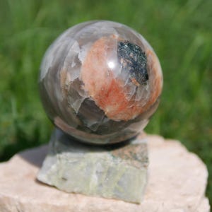Pode incluir: Uma pedra esférica polida com uma base cinzenta translúcida, uma inclusão proeminente de mineral cor de pêssego e uma mancha escura de aspeto metálico. A esfera repousa sobre uma base de pedra retangular, rugosa e de cor clara.