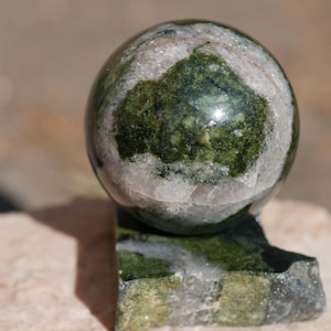Puede incluir: Una piedra esférica pulida con patrones cristalinos de color verde bosque profundo y blanco translúcido. La esfera descansa sobre una base de piedra rectangular a juego con inclusiones minerales verdes y blancas y textura rugosa.