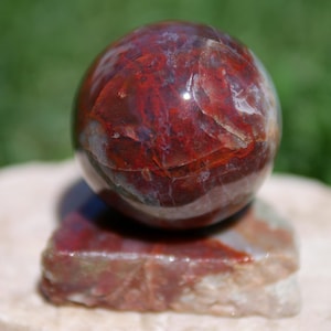 Peut inclure: Une sphère polie, aux tons rouges et bruns, posée sur un socle carré de même matériau. L'orbite présente un aspect marbré avec des motifs tourbillonnants. L'arrière-plan est vert flou, suggérant un cadre extérieur. Objet décoratif.