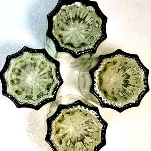 May include: Four clear glass tumblers with a starburst design and black rims are arranged in a cross formation. The glasses have a textured interior and are set against a white background, creating a striking contrast.