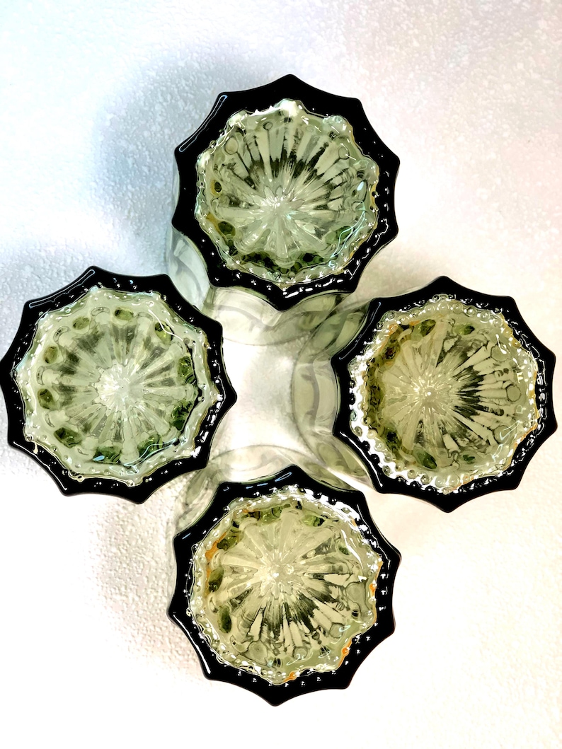 May include: Four clear glass tumblers with a starburst design and black rims are arranged in a cross formation. The glasses have a textured interior and are set against a white background, creating a striking contrast.