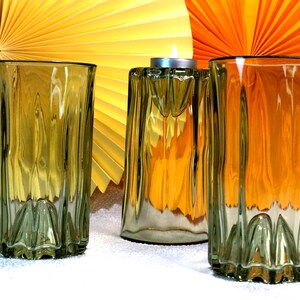 May include: Three olive-green glass items: two tall, ribbed drinking glasses and a candle holder with a lit tealight. The background features yellow and orange paper fan decorations. The glasses have a textured, ridged design.