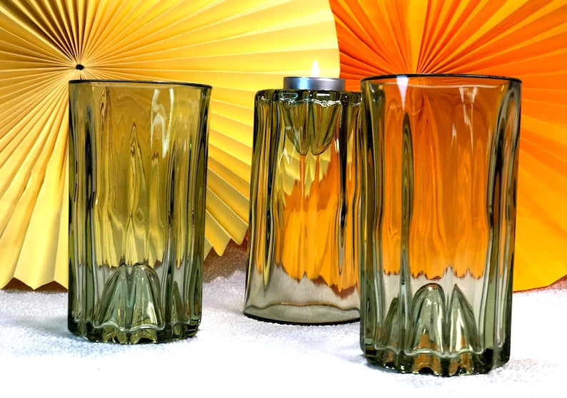 May include: Three olive-green glass items: two tall, ribbed drinking glasses and a candle holder with a lit tealight. The background features yellow and orange paper fan decorations. The glasses have a textured, ridged design.
