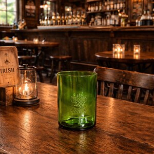 May include: A green glass tumbler with a tree design etched on the front sits on a wooden table in a pub. The table is surrounded by wooden chairs and tables. A lit candle is in a glass lantern.