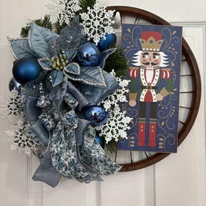 May include: A Christmas wreath with a wooden wheel base. The wreath features blue ornaments, snowflakes, and a large blue bow. A rectangular sign with a nutcracker illustration is attached to the wheel.