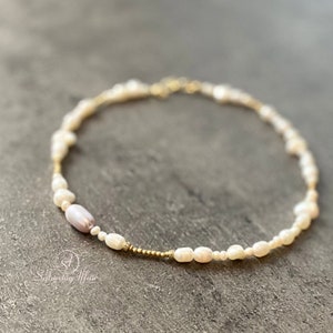 Könnte beinhalten: Zartes Perlenarmband mit einer Mischung aus kleinen, unregelmäßigen weißen Perlen und einer einzelnen, größeren, lavendelfarbenen Perle. Das Armband ist an einer goldfarbenen Kette und einem Verschluss befestigt. Das Logo "Saturday Affair" ist unten links sichtbar.
